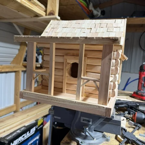 A log cabin birdhouse on the bench with its shingle roof going on