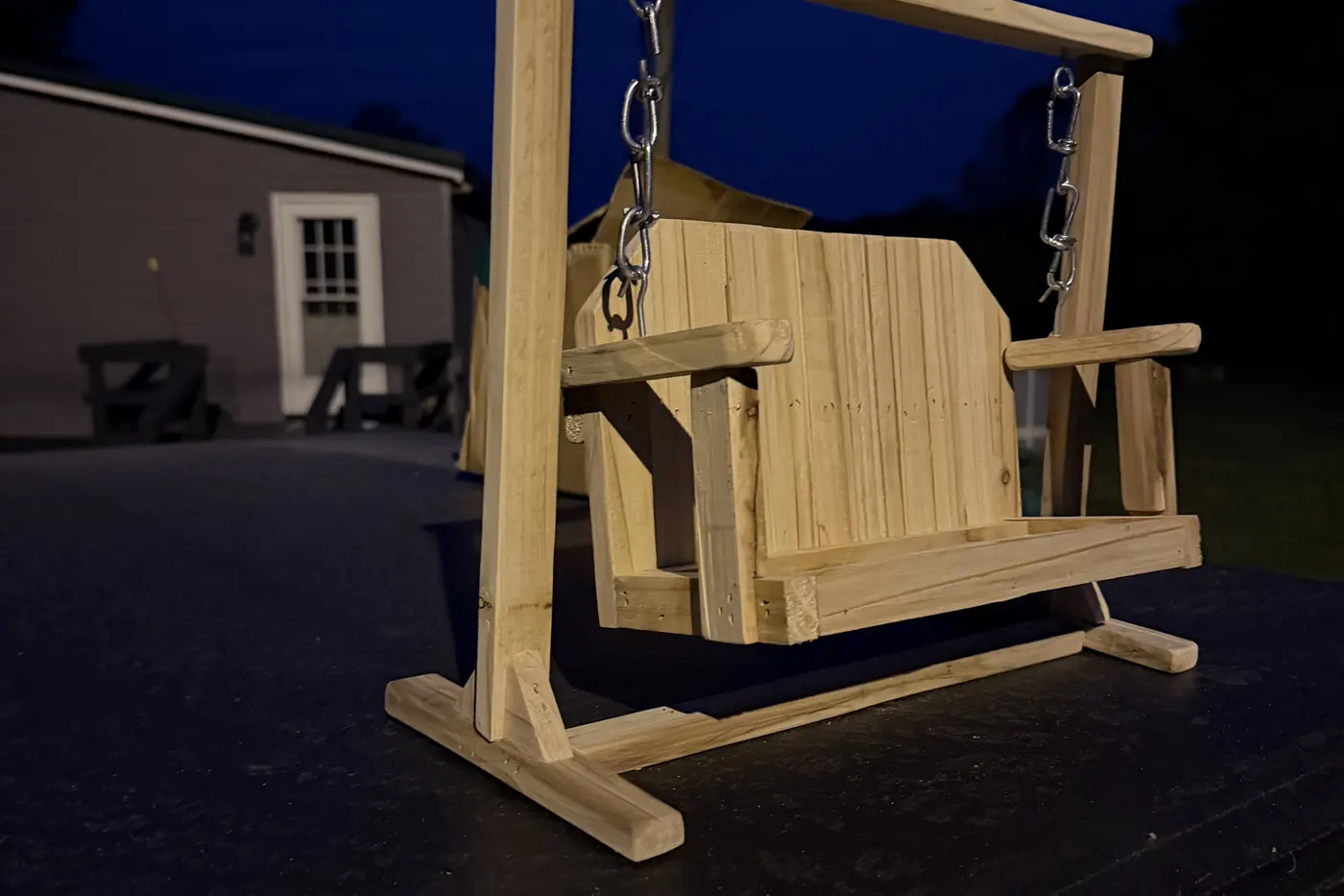 A full-size cedar porch swing hanging from its own A-frame stand on a deck at dusk.