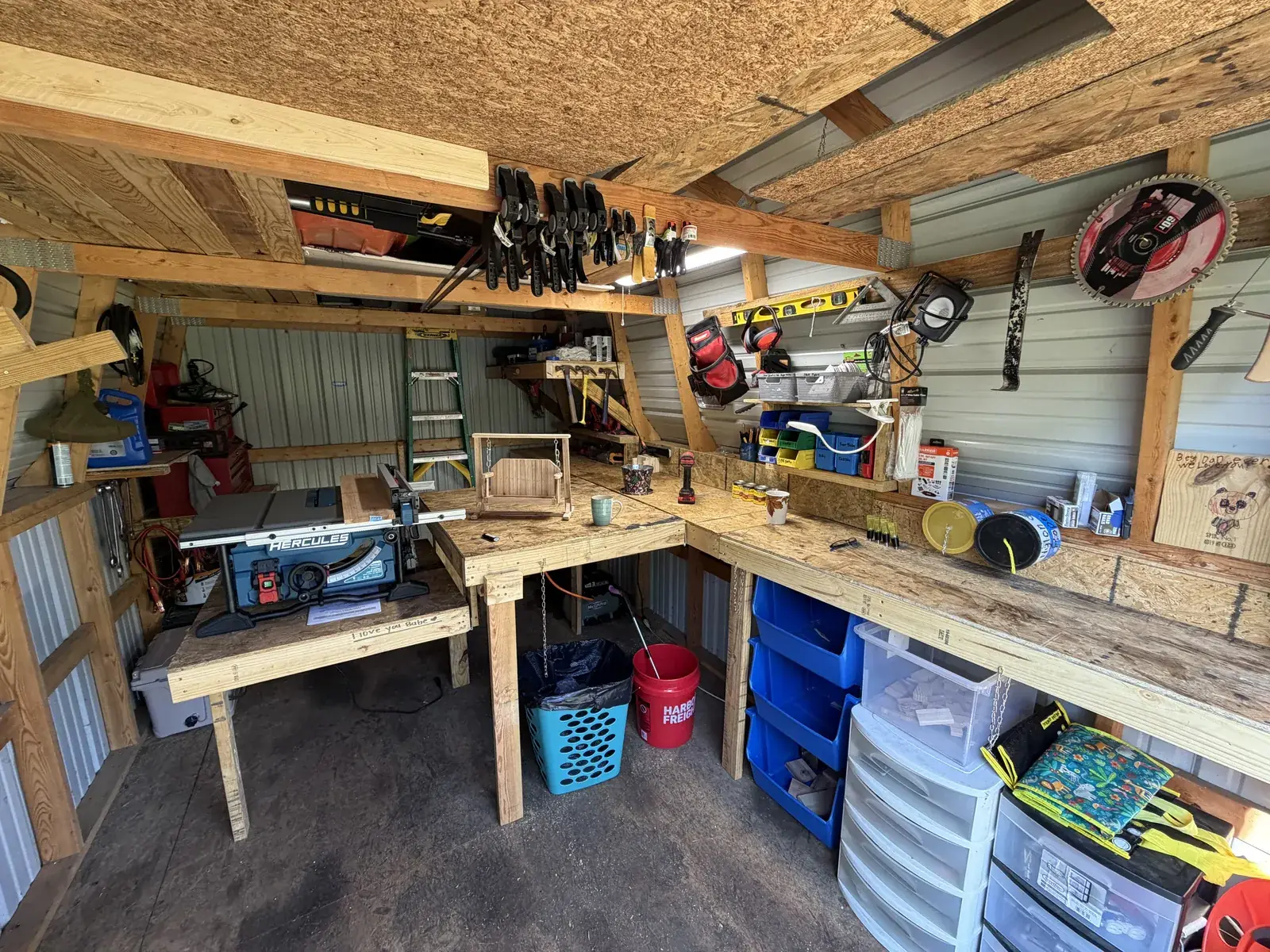 The corner of Harley's shop: table saw, a rail of clamps overhead, storage bins, and a swing sat on the bench mid-build
